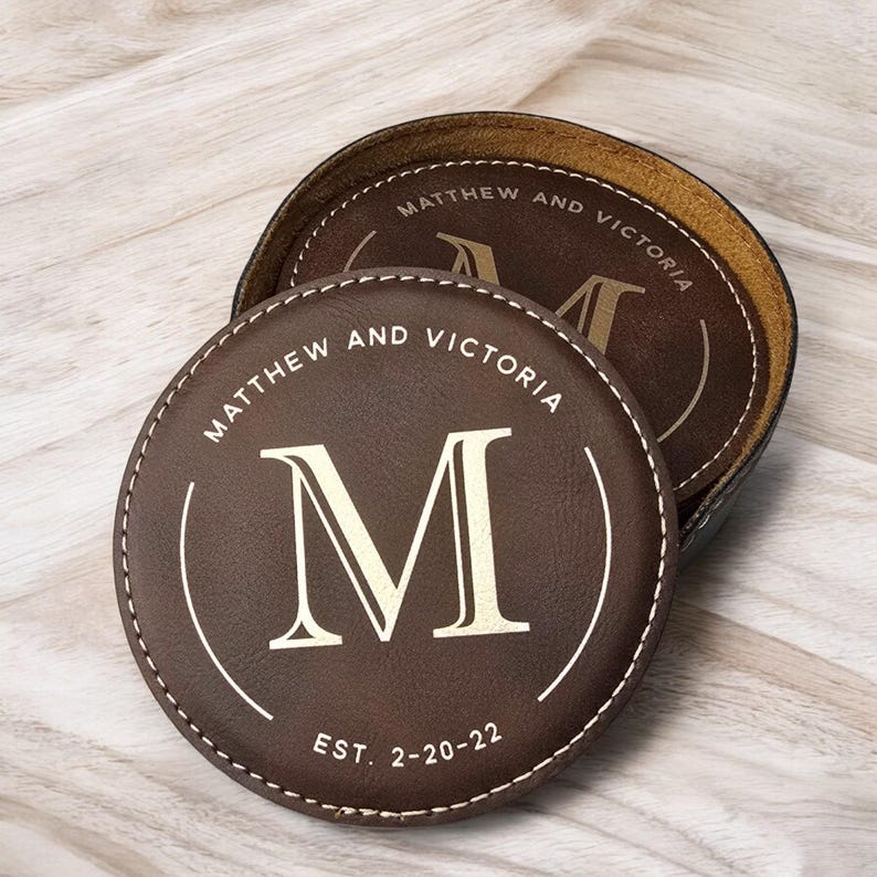K&ouml;nnte beinhalten: Zwei braune Lederuntersetzer mit gen&auml;hten Kanten. Die Untersetzer zeigen ein goldenes Monogramm "M" und den Text "Matthew und Victoria Est. 2-20-22".