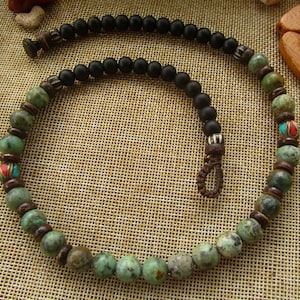 Pode incluir: Um colar de contas verdes e pretas com um fecho de corda castanho. O colar apresenta contas redondas verdes com detalhes castanhos e algumas contas vermelhas.