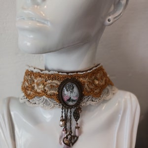 Peut inclure: Un collier ras-du-cou composé d'une large bande de dentelle superposée dorée et crème. Le centre est orné d'un pendentif camée ovale représentant une silhouette avec des fleurs roses, agrémenté de chaînes en bronze, perles et perles roses.
