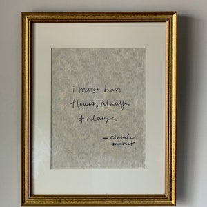 Może przedstawiać: Wydruk w ramie z cytatem Claude'a Moneta, który brzmi: "Muszę mieć kwiaty zawsze i zawsze". Wydruk jest na teksturowanym, kremowym papierze i jest oprawiony w złotą ramę.