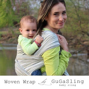 May include: A woman wearing a green shirt is carrying a baby in a woven baby wrap sling. The sling is a striped pattern of white, gray, and green. The baby is wearing a light green shirt. The woman and baby are outdoors in a natural setting.