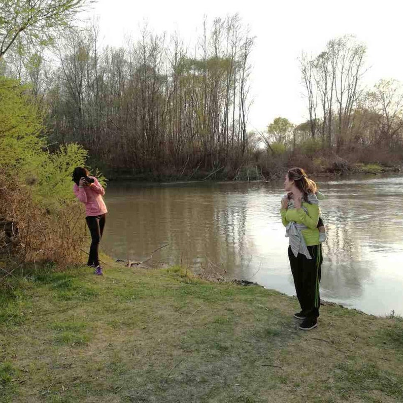 May include: Two people standing by a river. One person is taking a photo while the other person is looking away from the camera.