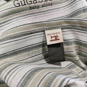 May include: A white and green striped baby sling with the text "GuGaSling baby sling" and a brown tag that says "Handmade" with a sewing machine icon.