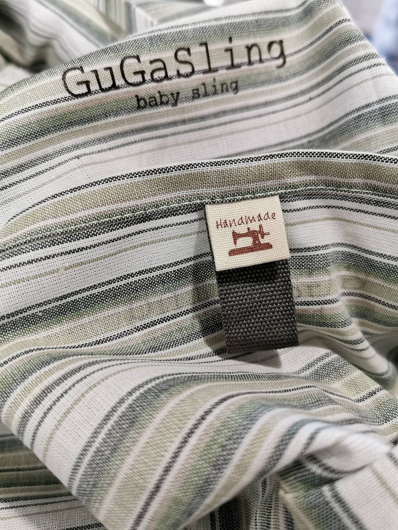 May include: A white and green striped baby sling with the text "GuGaSling baby sling" and a brown tag that says "Handmade" with a sewing machine icon.