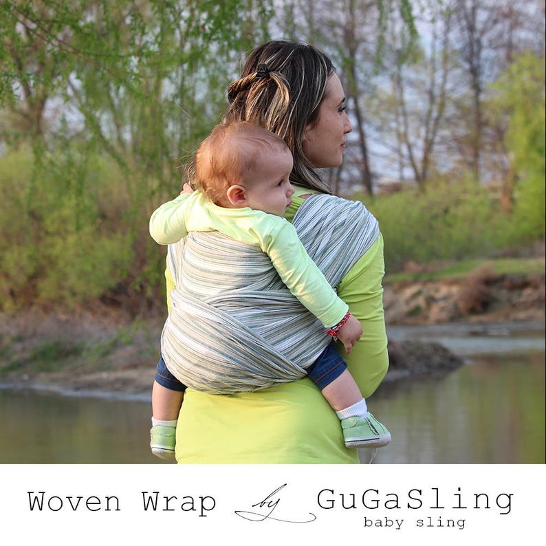 May include: A woman wearing a green shirt is carrying a baby in a woven baby wrap with a striped pattern. The wrap is a light gray and green color. The baby is wearing a yellow shirt and has a green and white striped baby sling. The text "Woven Wrap by GuGaSling baby sling" is at the bottom of the image.