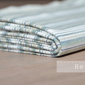 May include: A folded fabric with a white background and green, blue, and beige stripes. The fabric is folded in a neat stack and the word "Herb" is written in white text on a white background.