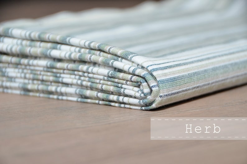 May include: A folded fabric with a white background and green, blue, and beige stripes. The fabric is folded in a neat stack and the word "Herb" is written in white text on a white background.