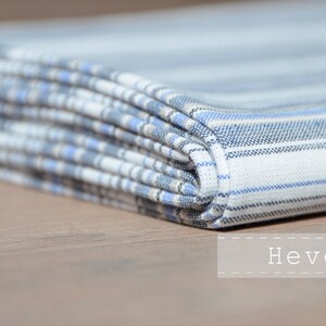 May include: A folded white and blue striped fabric with a subtle plaid pattern. The fabric is folded in a neat stack and the word "Heven" is printed below the fabric.