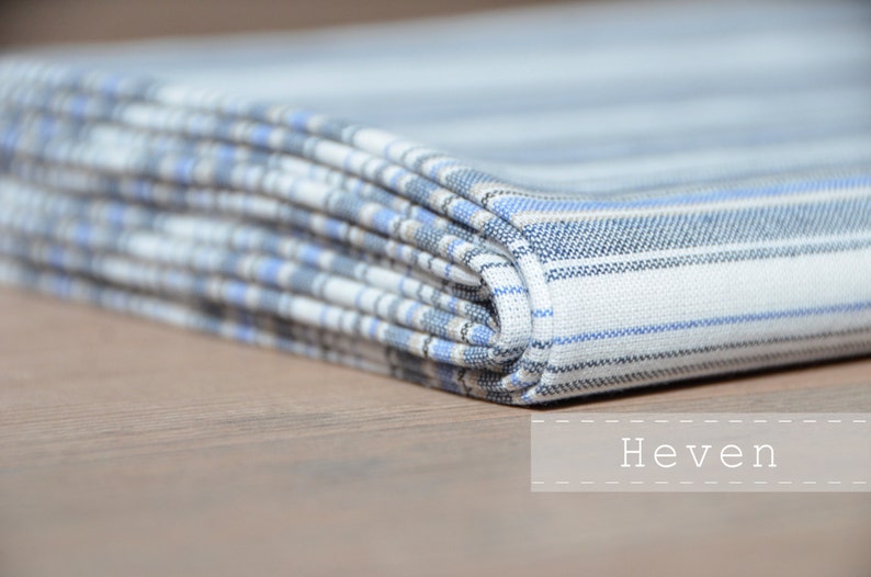 May include: A folded white and blue striped fabric with a subtle plaid pattern. The fabric is folded in a neat stack and the word "Heven" is printed below the fabric.