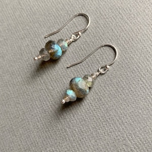 May include: A pair of handcrafted earrings featuring faceted, iridescent blue-green and gray beads. The beads are strung on a silver-colored wire, with a simple hook design. The earrings are displayed on a gray surface.