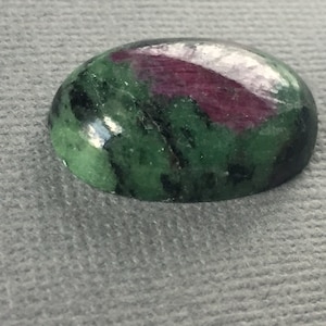 May include: Oval-shaped green and pink gemstone with black inclusions. The gemstone is polished and has a smooth surface.