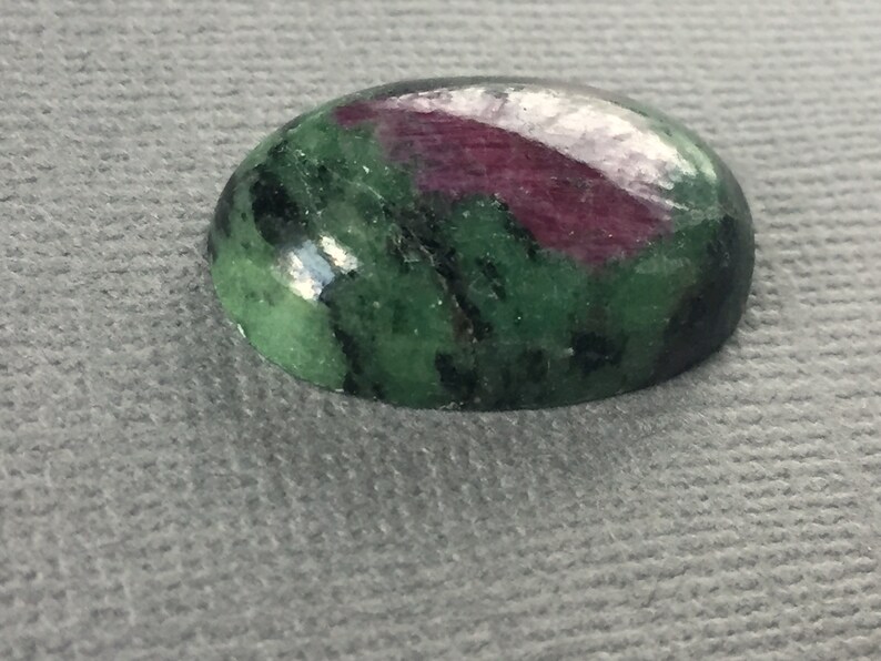 May include: Oval-shaped green and pink gemstone with black inclusions. The gemstone is polished and has a smooth surface.