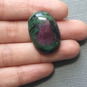 May include: Oval-shaped green and pink gemstone with black inclusions. The gemstone is cabochon cut and has a smooth, polished surface.