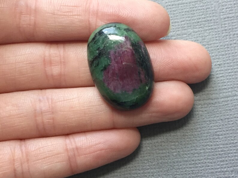 May include: Oval-shaped green and pink gemstone with black inclusions. The gemstone is cabochon cut and has a smooth, polished surface.