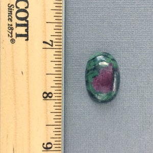 May include: Oval-shaped green and pink gemstone cabochon with a ruler for size reference. The gemstone is approximately 1/2 inch in length.