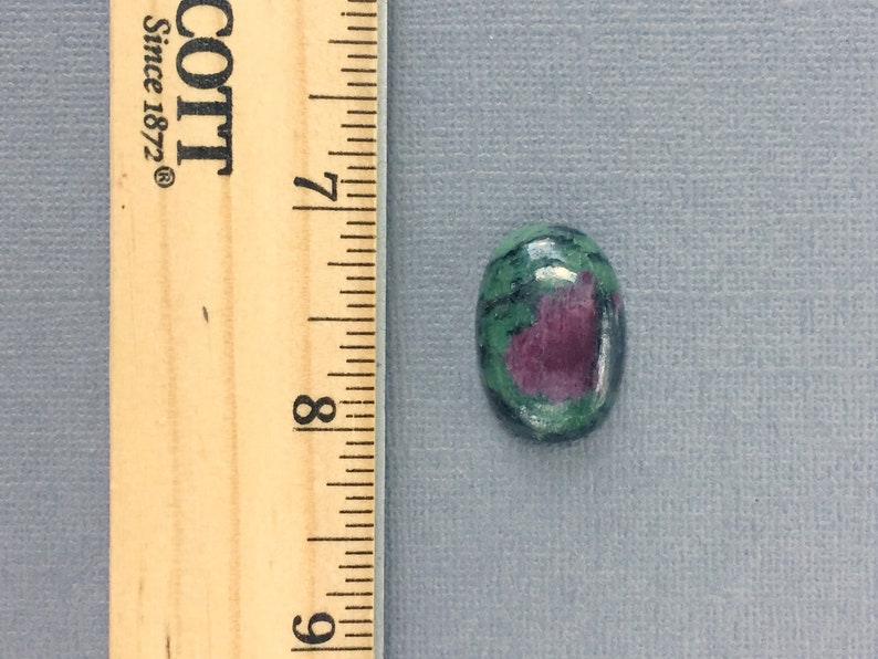 May include: Oval-shaped green and pink gemstone cabochon with a ruler for size reference. The gemstone is approximately 1/2 inch in length.