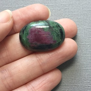 May include: Oval-shaped green and pink gemstone with black veins. The gemstone is cabochon cut and has a smooth, polished surface.