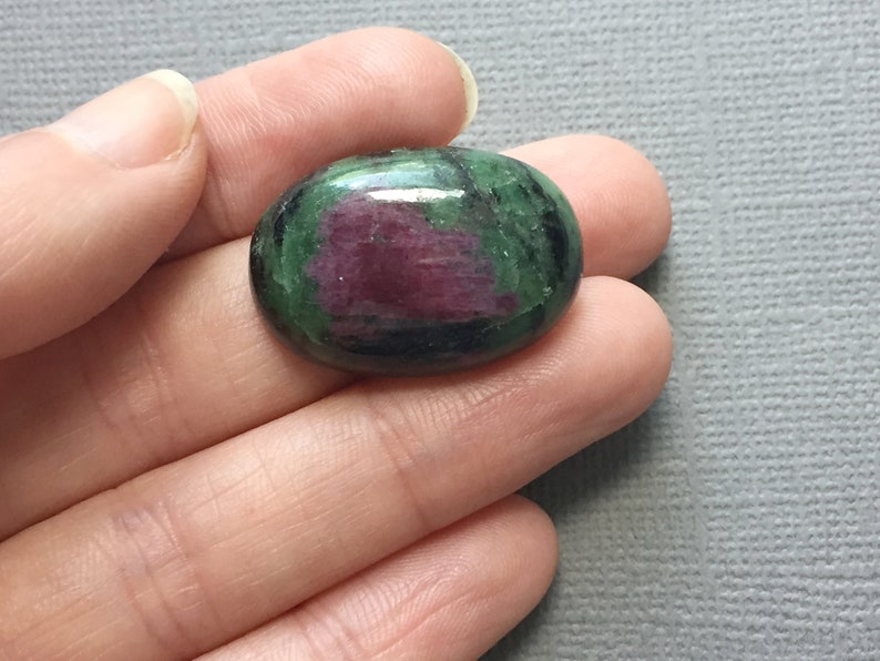 May include: Oval-shaped green and pink gemstone with black veins. The gemstone is cabochon cut and has a smooth, polished surface.