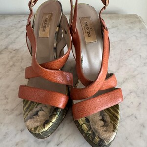 May include: A pair of orange leather Valentino high-heeled sandals. The sandals feature crisscross straps and ankle straps with gold buckles. The footbeds have a floral pattern. The heels are approximately 4 inches high.
