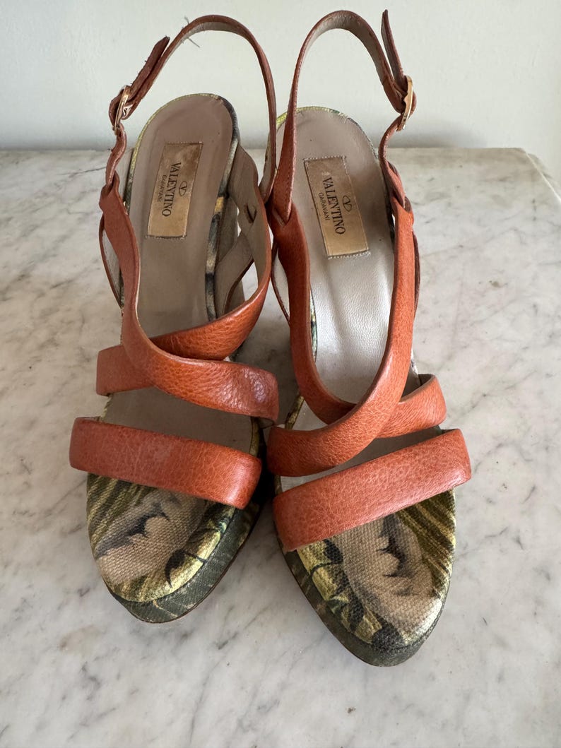 May include: A pair of orange leather Valentino high-heeled sandals. The sandals feature crisscross straps and ankle straps with gold buckles. The footbeds have a floral pattern. The heels are approximately 4 inches high.