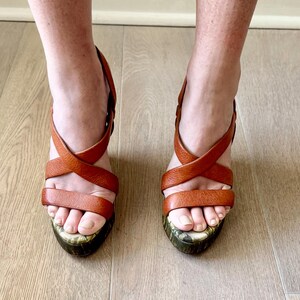 May include: A pair of brown leather platform sandals with crisscross straps. The sandals have a patterned sole and are worn on a light wood floor. The shoes are a warm, earthy tone.