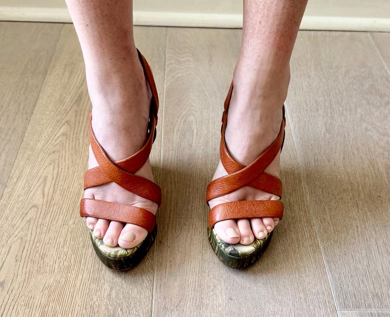 May include: A pair of brown leather platform sandals with crisscross straps. The sandals have a patterned sole and are worn on a light wood floor. The shoes are a warm, earthy tone.