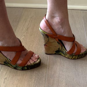 May include: Brown leather wedge sandals with multiple straps. The wedge heel features a floral pattern with green and yellow accents. The sandals have a buckle closure and are worn on a light wood floor.