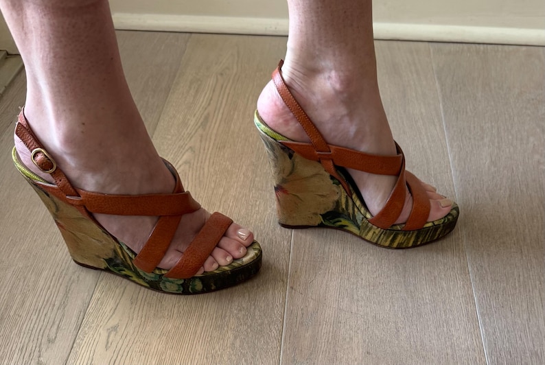 May include: Brown leather wedge sandals with multiple straps. The wedge heel features a floral pattern with green and yellow accents. The sandals have a buckle closure and are worn on a light wood floor.
