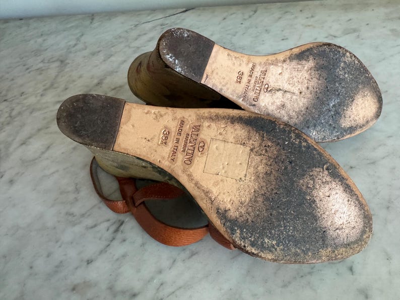 May include: A pair of brown leather Valentino sandals, size 38, with a wedge heel. The soles are worn and show signs of use. The sandals have a strap design and are made in Italy.