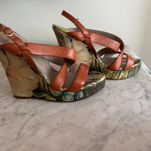 May include: A pair of wedge sandals with orange leather straps and a floral print fabric. The heels are approximately 4 inches high. The sandals have a buckle closure and a patterned sole.