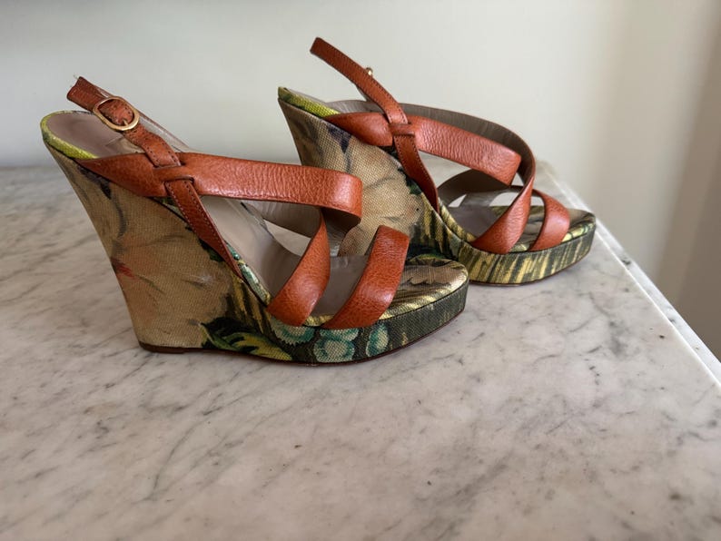 May include: A pair of wedge sandals with orange leather straps and a floral print fabric. The heels are approximately 4 inches high. The sandals have a buckle closure and a patterned sole.
