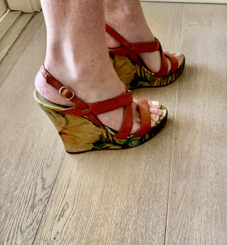May include: Red leather wedge sandals with floral print platforms. The sandals feature multiple straps across the foot and ankle straps with gold buckles. The platform has a colorful floral design.