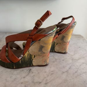 May include: A pair of wedge sandals with brown leather straps and a floral patterned fabric on the wedge heels. The sandals have a buckle closure and a multi-colored floral design.