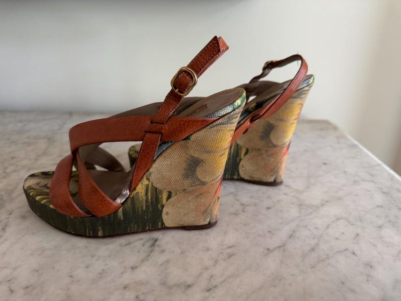 May include: A pair of wedge sandals with brown leather straps and a floral patterned fabric on the wedge heels. The sandals have a buckle closure and a multi-colored floral design.