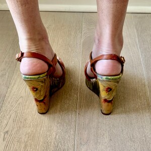 May include: Brown leather wedge sandals with floral print on the wedge heel. The sandals have a strap around the ankle and a strap across the toes. The heels are approximately 4 inches high.