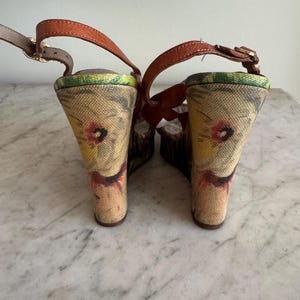 May include: Pair of wedge sandals with brown leather straps and floral print fabric heels. The heels have a wedge design and are approximately 4 inches (10 cm) high. The sandals have a buckle closure and a neutral color palette.