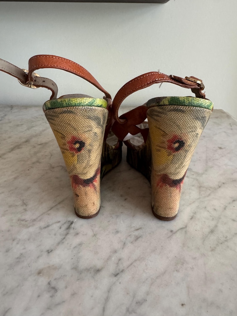 May include: Pair of wedge sandals with brown leather straps and floral print fabric heels. The heels have a wedge design and are approximately 4 inches (10 cm) high. The sandals have a buckle closure and a neutral color palette.