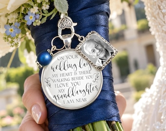 Puede incluir: Un dije plateado para ramo de novia que incluye un colgante redondo con el texto ON YOUR WEDDING DAY MY HEART IS THERE WALKING BESIDE YOU I LOVE YOU MY GRANDDAUGHTER I AM NEAR, un marco de fotos cuadrado y una cuenta azul.