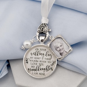 May include: A silver wedding memorial charm with a photo and text. The charm has a round pendant with the words "ON YOUR wedding day... I LOVE YOU my granddaughter I AM NEAR." A small photo frame is attached. A white ribbon is at the top.