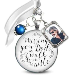 May include: A white ribbon with a silver charm that says "Missing you Dad as I walk down the aisle". A small blue bead and a silver frame with a photo of a man are attached to the charm.
