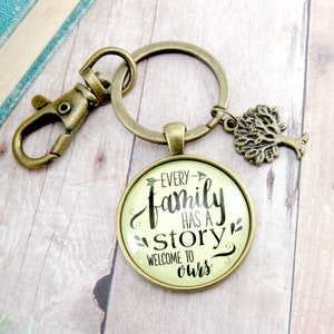 May include: A bronze-coloured keyring with a round pendant inscribed with the words "Every family has a story, welcome to ours." The keyring features a tree charm and a clasp. The text is in black script on a cream background.