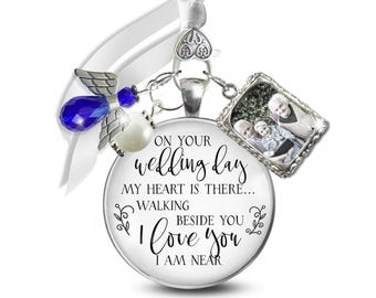May include: A silver-toned memorial charm featuring a round pendant with the text "ON YOUR wedding day MY HEART IS THERE... WALKING BESIDE YOU I LOVE YOU I AM NEAR". It includes a blue crystal angel wing charm, a white pearl, and a rectangular photo frame.