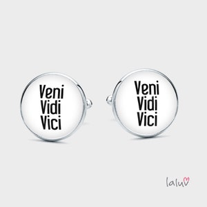 Op de afbeelding: Een paar zilveren manchetknopen met witte ronde vlakken. De vlakken zijn bedrukt met de Latijnse zin "Veni Vidi Vici" in zwart.