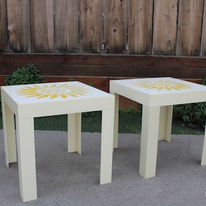 May include: Two white end tables with yellow floral designs on the top. The tables have a square top and four legs.