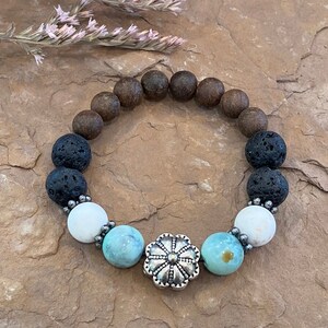May include: A beaded bracelet featuring a mix of natural stone beads. The bracelet includes dark brown, black lava, white, and turquoise-colored beads. A silver-colored flower-shaped bead is the focal point of the bracelet. The bracelet is displayed on a textured, brown surface.