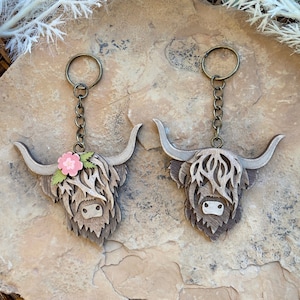 May include: Two wooden Highland Cow keychains with brown fur and black noses. One keychain has a pink flower on its head. Both keychains are attached to gold-colored chains.
