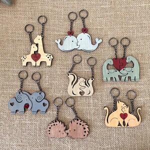 May include: Set of eight wooden keychains featuring different animal couples, each with a heart-shaped puzzle piece. The animals include whales, giraffes, squirrels, elephants, dinosaurs, cats, and hedgehogs. The keychains are made of natural wood and have a rustic, handmade look.