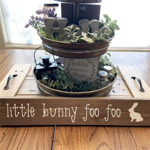 May include: A tiered tray with a rustic wooden base, featuring a sign that reads "little bunny foo foo". The tray is decorated with greenery, a lantern, and a sign that says "grateful thankful blessed".