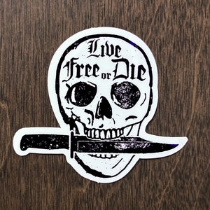 May include: Black and white sticker of a skull with a knife in its mouth. The skull has the words "Live Free or Die" written on it.