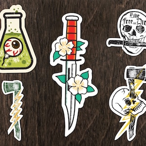 Può includere: Cinque adesivi in stile vintage con vari disegni. Un becher verde con un occhio all'interno, un pugnale rosso e bianco con fiori, uno scheletro con un coltello in bocca e il testo "Live Free or Die", un'ascia verde con un fulmine e uno scheletro con un fulmine.